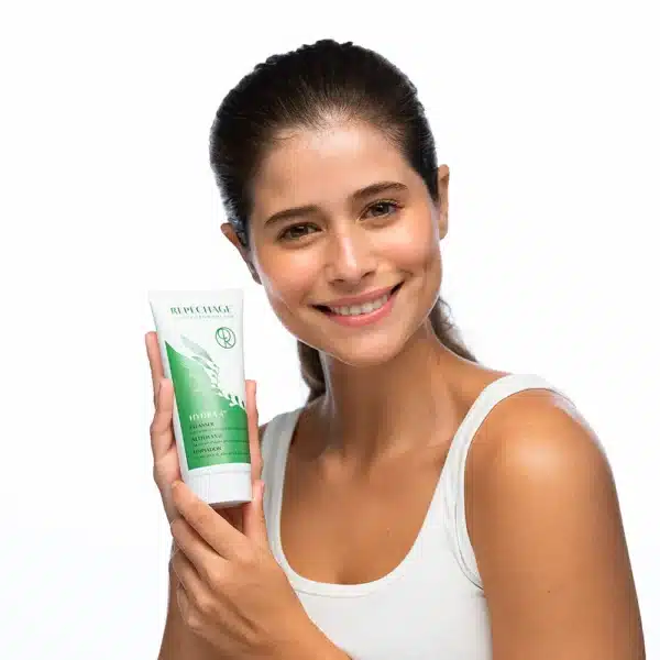 Nainen hymyilee ja pitää kädessään Repechage Hydra 4 Cleanser -kasvojenpuhdistusainetta, vihreä ja valkoinen tuubipakkaus.