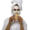 Nainen valkoisessa kylpytakissa ja pyyhe turbaanina, kasvoillaan valkoinen kasvonaamio ja kädessään hunajakennopala.