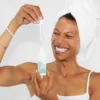 Nainen pyyhkeellä päähän kiedottuna käyttää pipettiä levittääkseen Repechage ihonhoitotuotetta pullosta, hymyillen iloisesti.